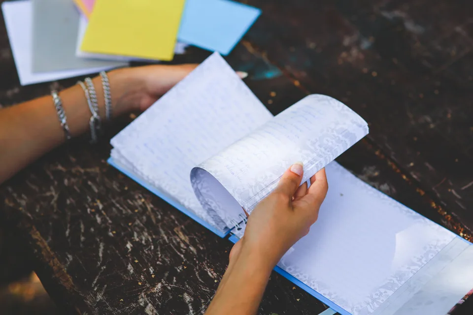 Hands turning a page in a handwritten notebook beside colored notes. Photo by Karolina Grabowska.